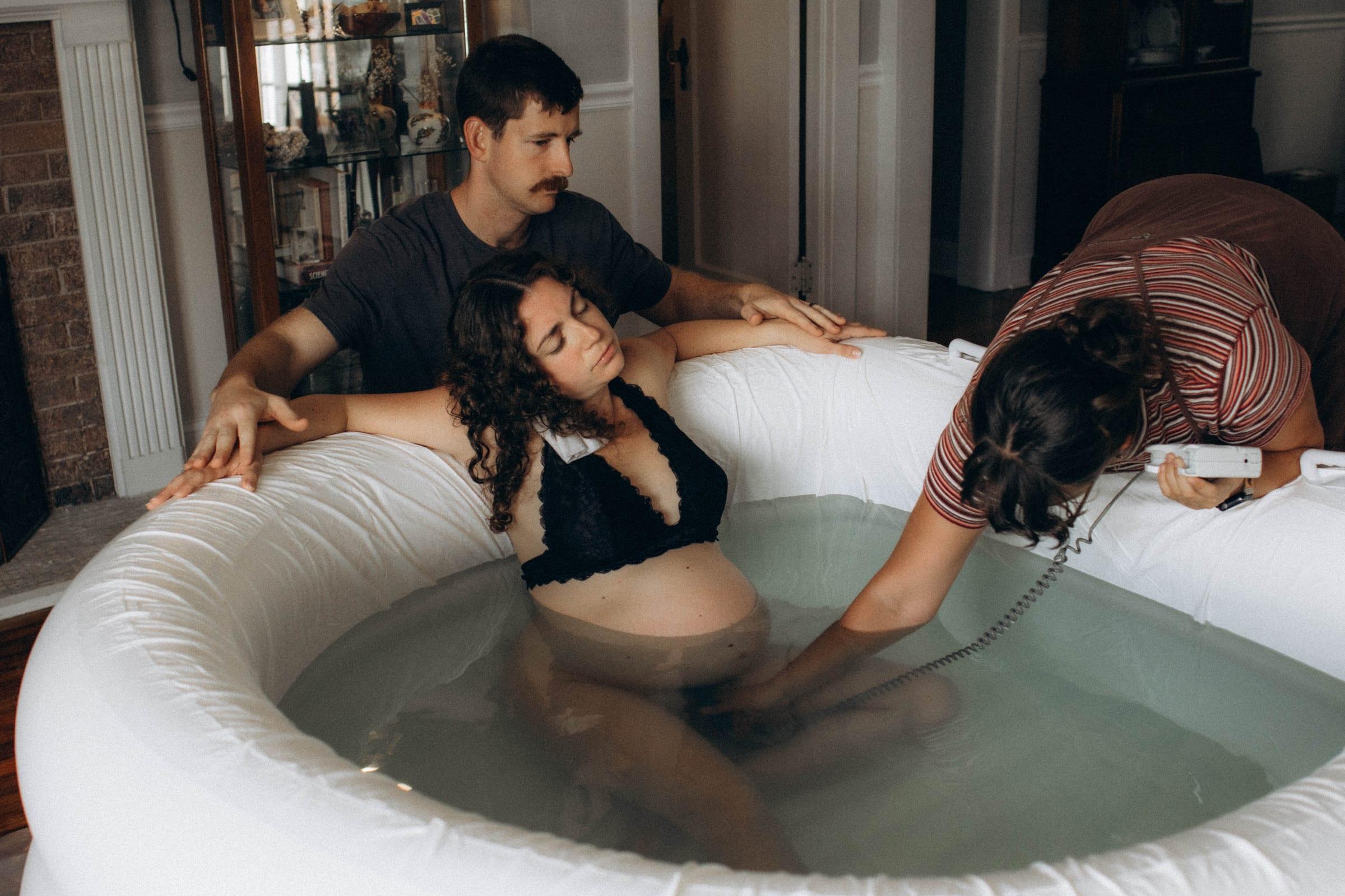 A mother laboring in a birth pool at home, her partner supporting her shoulders while a midwife listens to the baby with a waterproof doppler