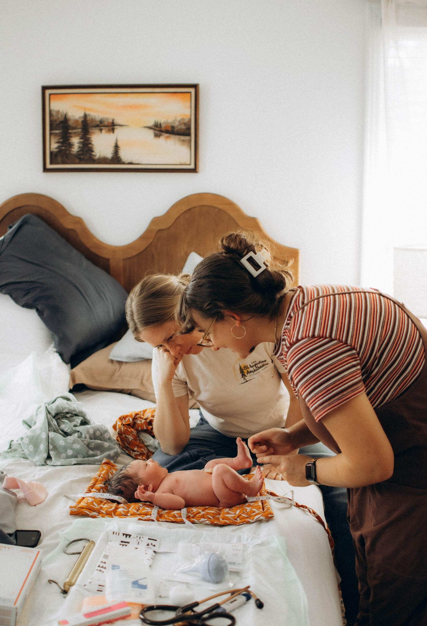 Two midwives leaning over a newborn on a bed during a newborn exam, their supplies laid out beside them